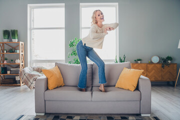 Cheerful blonde woman enjoying an energetic moment standing on a cozy sofa in a stylish and well...