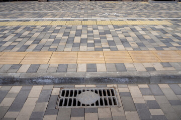 Modern sidewalk covered with decorative and tactile tiles for the visually impaired, as well as a sewage manhole on the roadway