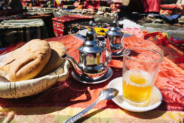 Traditional mint tea. Moroccan drink moment. Silver teapot shine. Fresh bread breakfast. River picnic vibes. Cultural table decor. Outdoor feast joy. Sunny day tea. Oriental hospitality.