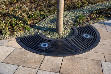 Semi-circular metal grating provides protection for tree roots. Stone mulch retains moisture. Light bulbs in grates for nighttime illumination. Aesthetic functionality in modern urban landscapes