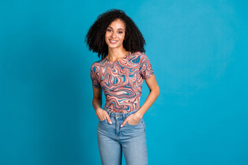 Stylish young woman with curly hair in a printed t-shirt on a vibrant blue background, expressing...