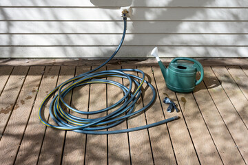 Coiled up garden hose, connected to spigot, laying on manufactured wood deck, with nozzle sprayer and plastic watering can  © knelson20