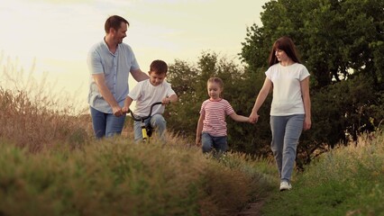 Mother holds running girl and father supports boy riding bicycle on family vacation at country area. Positive father helps boy on bicycle and mother holds active girl hand on family weekend at sunset