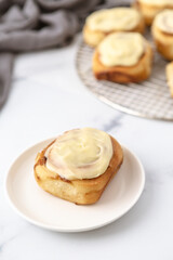 Homemade GLazed Cinnamon Roll on a White Plate and White Marble Kitchen Counter; Additional Rolls on Cooling Rack in Background