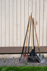 Spring gardening, two shovels and a rake leaning against a light brown sided exterior house wall,...