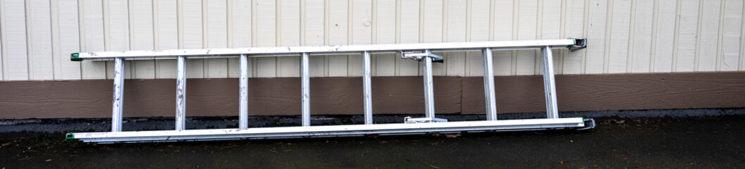 Aluminum extension ladder laying on asphalt driveway against light brown wood siding exterior home wall  © knelson20