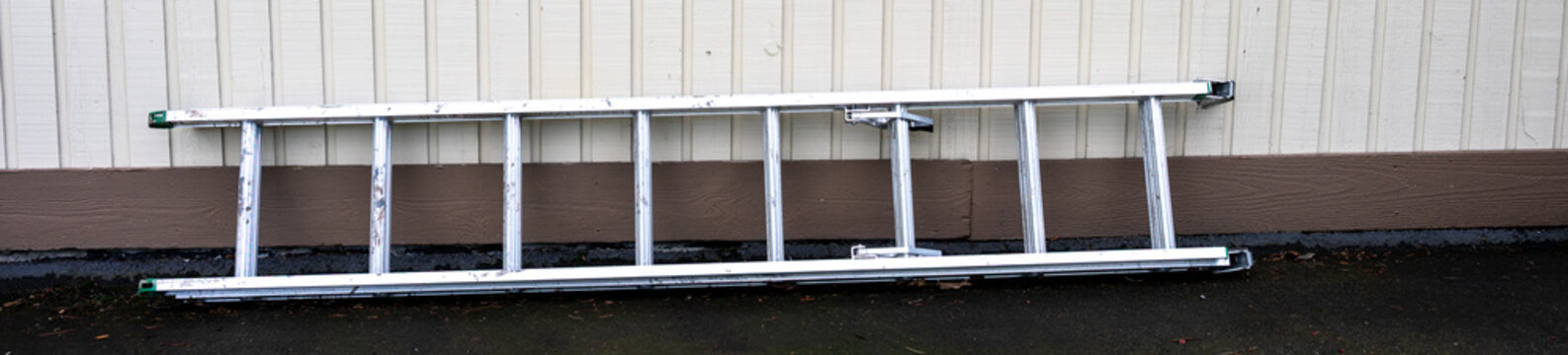Aluminum extension ladder laying on asphalt driveway against light brown wood siding exterior home wall
