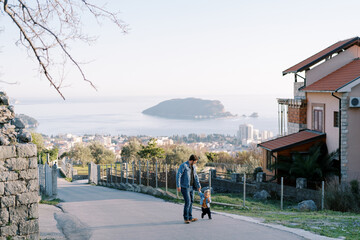 Naklejka premium Dad and a little girl walk holding hands along the road in a town above the sea