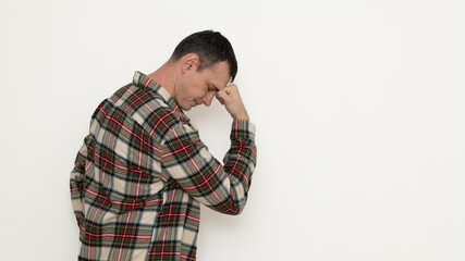 A man wearing a plaid shirt stands facing a plain wall, resting his forehead on his hand in a...