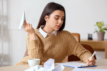 Young woman with paper plane writing at table in office