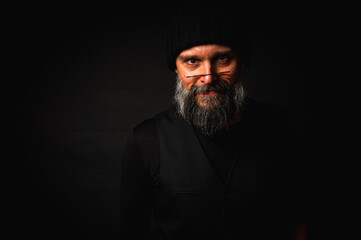 Mature man with a gray beard wearing round glasses, a black knitted hat and a vest. Studio portrait with dramatic lighting and a dark background. Hipster style, thoughtful, serious. Low key