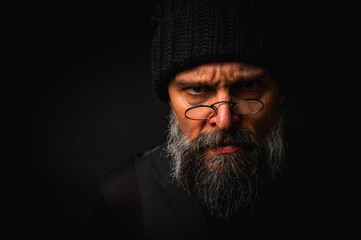 portrait, expressive mature man with a keen gaze and frown, wearing glasses and a knitted hat. Dramatic restrained portrait with strong emotions on a dark background