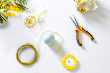Top view of Easter craft materials on a white table: gold wire, yellow ribbon, pliers, mimosa...