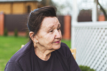 Elderly woman 70 80 years old with dark brown hair and earrings sitting outdoors near white lattice fence, looking to the side with calm thoughtful expression, concept aging, retirement, lifestyle