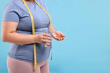 Plus size woman holding pile of weight loss supplements and glass with water on light blue...