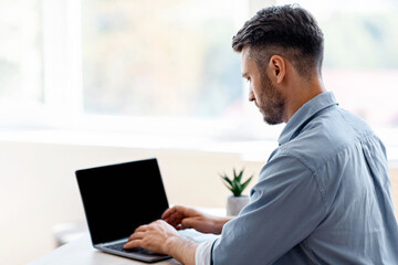 Fototapeta premium Busy Male Entrepreneur Working On Laptop With Black Blank Screen In Office, Typing On Keyboard, Over Shoulder View, Mockup