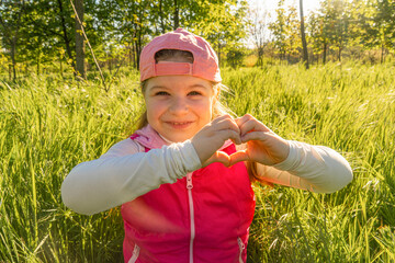 A cheerful girl is sitting in the thick green grass, depicting a heart sign with her hands. Walk outdoors in the city park