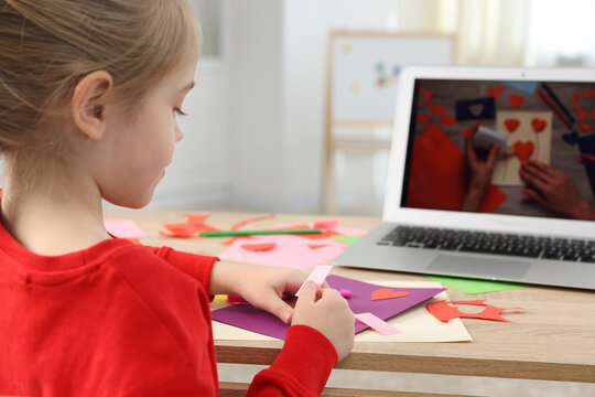 Little girl making handmade project with color paper by video tutorial at table indoors