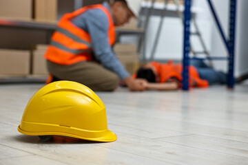Accident at work. Hardhat on floor and man checking pulse of his unconscious colleague in...