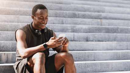 Sporty black guy using smartphone while sitting on urban stairs after training outdoors, resting after fitness workout in city park, free space