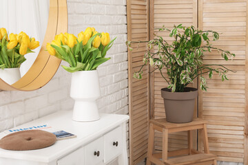 Vase of yellow tulips on dresser with mirror, dressing screen and houseplant near white brick wall...