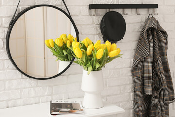 Vase of yellow tulips on dresser with mirror, hanger and coat near white brick wall in hallway
