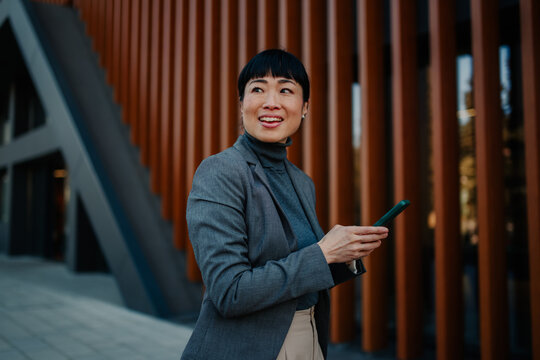 Businesswoman using smartphone and smiling near office building