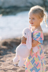 Little smiling girl with her hair fluttering in the wind stands on the beach with a plush bunny and...