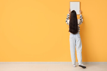 Young woman hanging picture frame on orange wall in room