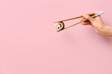 Hand with tasty sushi roll and chopsticks on pink background