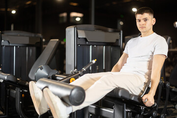 Focused athletic young man performing seated leg extensions on resistance machine, isolating...
