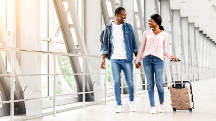 Fototapeta premium Journey Together. Smiling Black Spouses Walking With Luggage In Airport Terminal, Romantic African American Couple Holding Hands While Going To Gate, Having Honeymoon Or Vacation Trip, Free Space