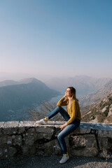 Naklejka premium Smiling girl sits on the curb of the observation deck over the Bay of Kotor. Montenegro