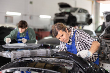 Focused experienced middle aged mechanic inspecting detached car bumper in workshop, scrutinizing damage, contemplating necessary steps for restoration or repair..