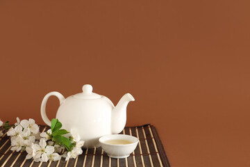 Teapot with cup of green tea and flowers on brown background