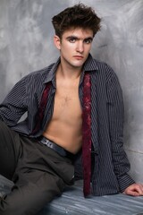 Young man with partially unbuttoned shirt and loosened tie, posing against a textured grey...