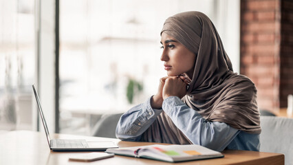 Desperate young muslim woman with laptop and notepad sitting at cafe, looking through the window,...