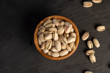 fried unpeeled pistachio nuts in a wooden bowl with cracked shells, closeup, top view