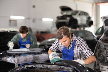 Adult male mechanic in uniform polishes part of car with machine in car service station