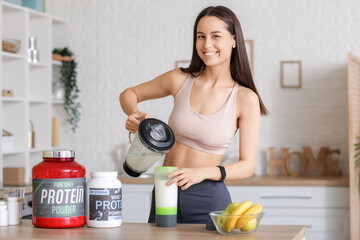 Sporty young happy woman with jars of protein powder and fruits pouring shake from jug into bottle ...