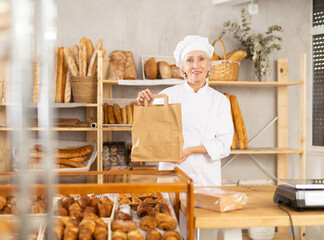 Portrait of helpful female baker with paper bag of fresh baked goods in interior of private bakery