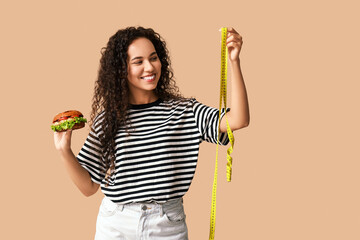 Happy young African-American woman with measuring tape and burger on beige background. Diet concept