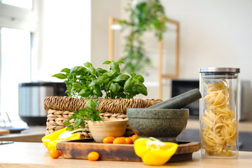 Basil plants with vegetables, pasta and mortar on table in kitchen