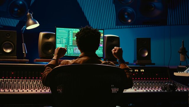 African american sound engineer feeling happy with his recorded song on mixing console, working in music industry post production. Audio technician making a hit song in control room. Camera B.