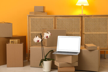 Blank laptop with orchid flower, commode and moving boxes near orange wall in room