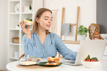 Young happy woman holding laptop, tasty toast with fresh butter and cheese in kitchen at home