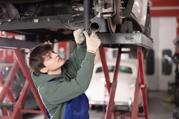 Young guy mechanic in uniform repairs underbody of car in car service station