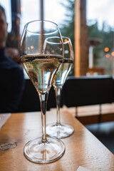 Two champagne flutes on a wooden table, reflecting warm light. Large windows reveal lush green...