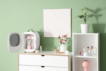 Open small refrigerator with cosmetic products and flowers in vase on chest of drawers near green wall in room. Closeup