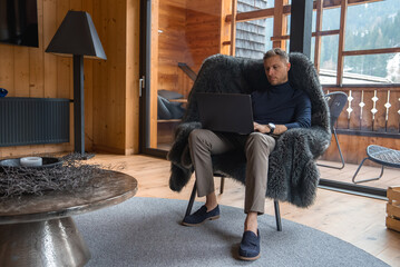 A man sits in a fur covered chair inside a wooden chalet with modern decor. Glass doors reveal a...